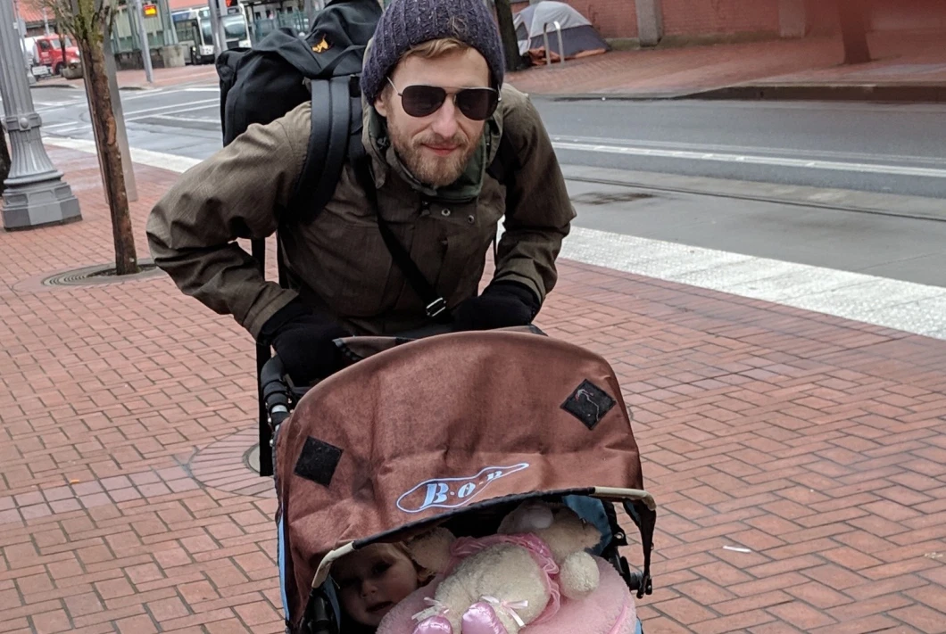 Andrew Anderson wearing a coat, gloves, beanie, and sunglasses and pushing a stroller containing a small child